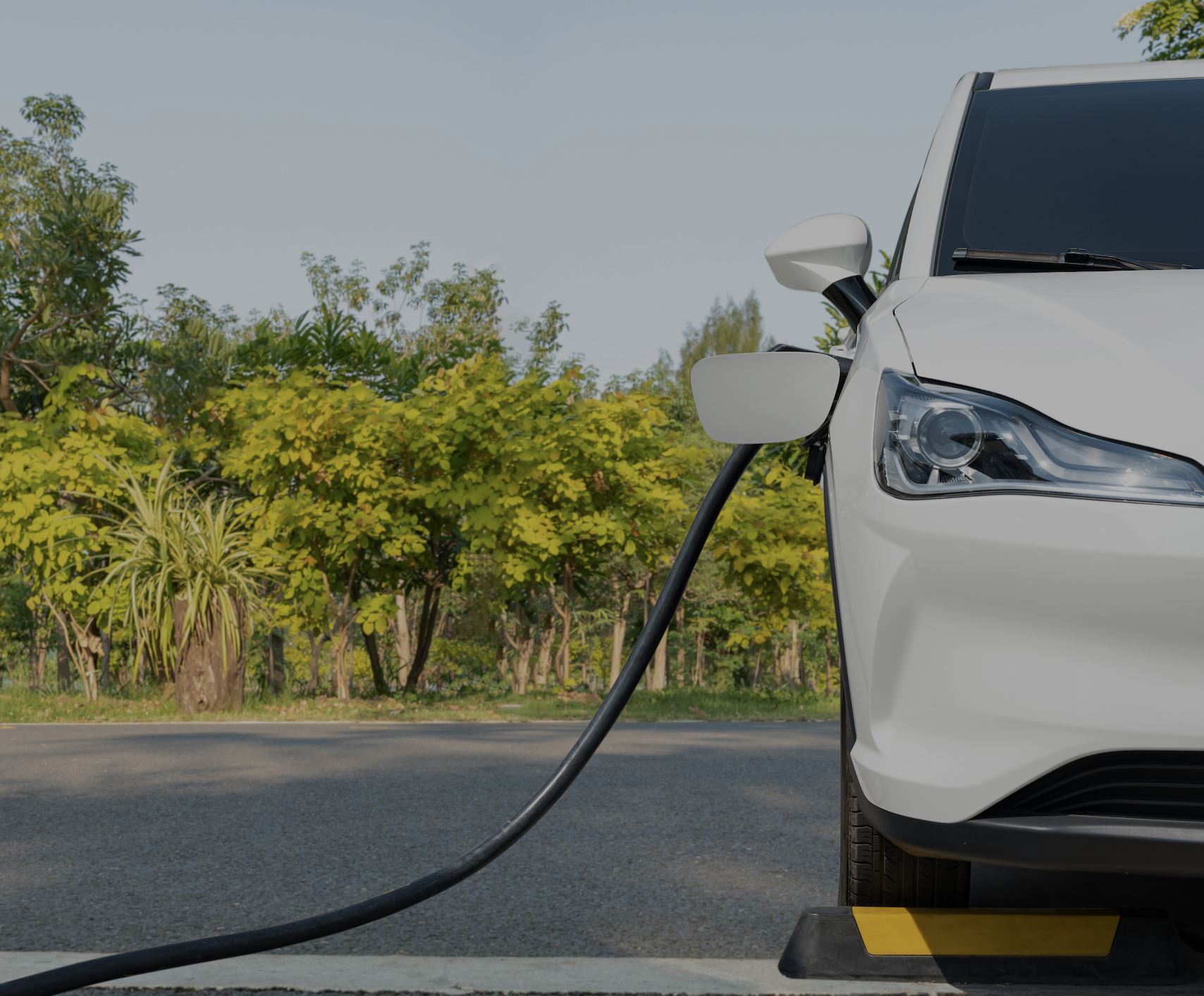 A white electric vehicle plugged into a charging station outdoors with trees in the background.
