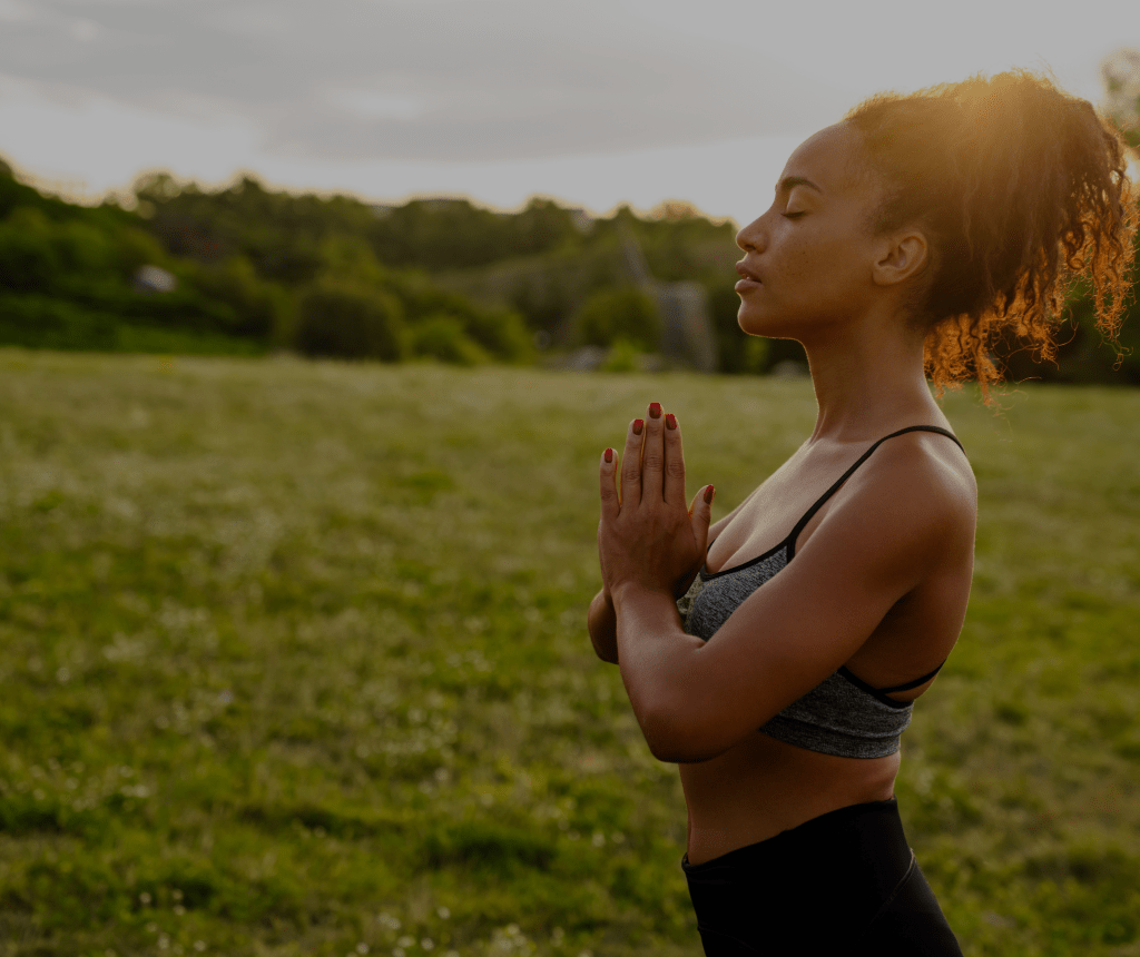 A side view of a woman with her hands clasped together standing in a serene field in a yoga pose.
